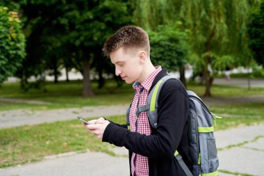 Kent parkta yürüyüş ve mobil akıllı telefon açık havada kullanma sırt çantası ile ekose gömlekli yakışıklı genç adam. Öğrenci ile smartphone, boş alan. Seyahat kavramı 