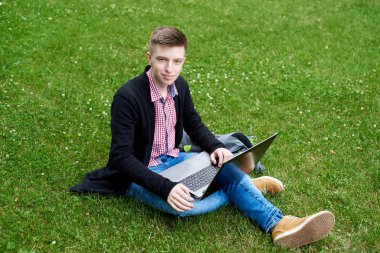 Okuma, dizüstü bilgisayarda çalışarak Yong adam ekose gömlek, ceket ve kot kampüste, yeşil çim üzerinde oturuyor. Öğrenci dizüstü bilgisayar açık havada, diz ile boş alan