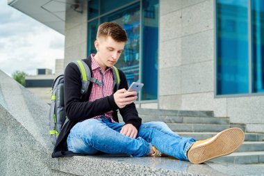 Yakışıklı delikanlı taş merdiven yanında oturan ve akıllı telefon şehir açık, tam vücut kullanarak sırt çantası ile. Üniversiteden sonra kopya alanı öğrenci aittir