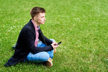 Doğa, kopya alanı yeşil çim çim üzerinde oturan ve mobil akıllı telefondan açık havada, kulaklık ile müzik dinlemek genç mutlu adam portresi