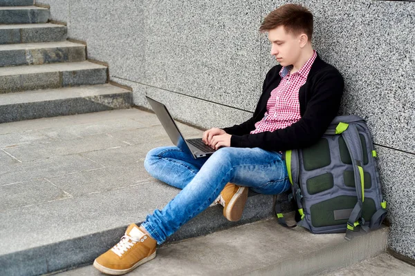 Handsome young man in jacket and jeans with backpack sitting on the stairs under grey stone wall and typing on laptop computer outdoors. Technology and communication concept