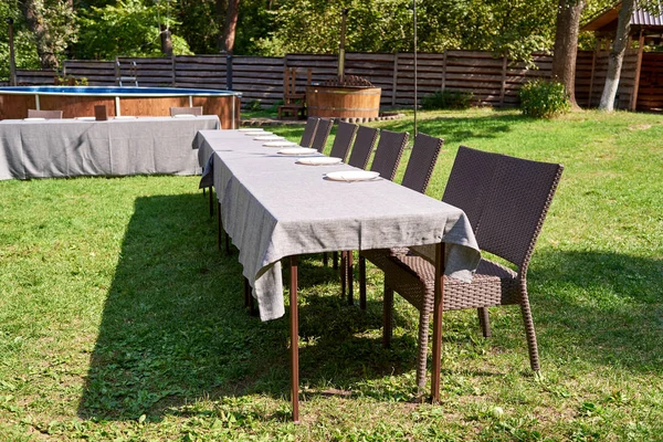 Dinner table with empty ceramic round plates and cutlery on grey tablecloth and chairs in garden outdoors, copy space. Dinner plate setting. White plate with knife and fork