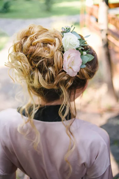 Beautiful bride in silk robe with fashion wedding hairstyle with fresh flowers, back view. Blond girl with curly hair styling. Hairstyle for long hair