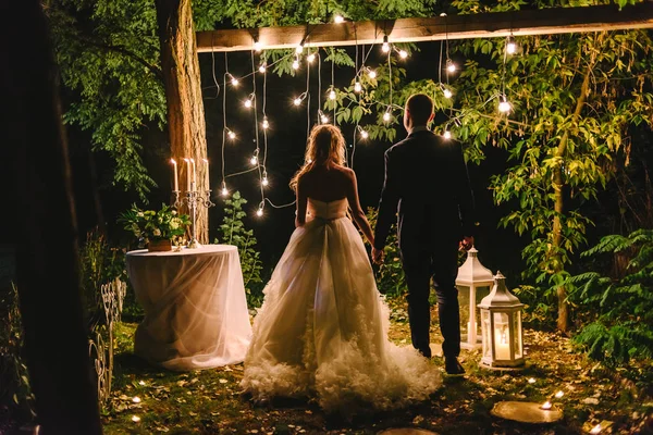 Night wedding ceremony with candles, lanterns and lamps on tree. Bride and groom holding hands on background of bulb lights, back view. Beautiful young couple standing under a tree at night