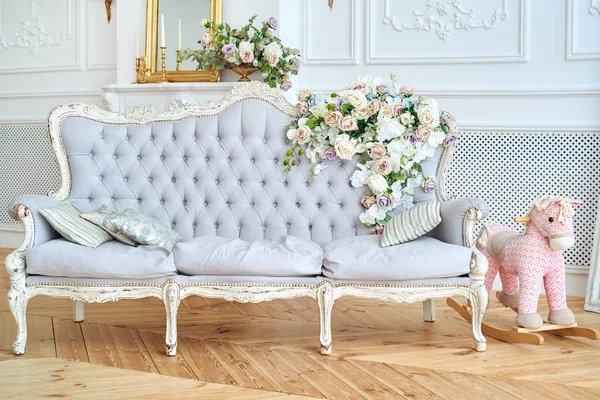 Soft sofa with gray fabric upholstery, pillows and flowers on it, copy space. Luxury rich living room interior design with elegant classic furniture, rocking horse, mirror and candles. Vintage textile couch