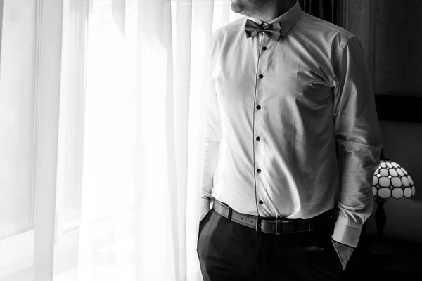 Man in white shirt, pants and bow tie standing at window indoors, close-up. Businessman dressing up. Fashion, style, lifestyle, groom