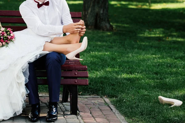 Wedding couple sitting on bench and relaxing in park outdoors, copy space. Groom doing foot massage to bride. Husband massaging his wife feet. Pain of wearing high heels