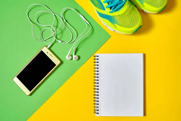 Top view of sport shoes, blank empty notebook and mobile cellphone with earphones on colorful background, copy space. Flat lay. Sport, fitness concept, healthy lifestyle