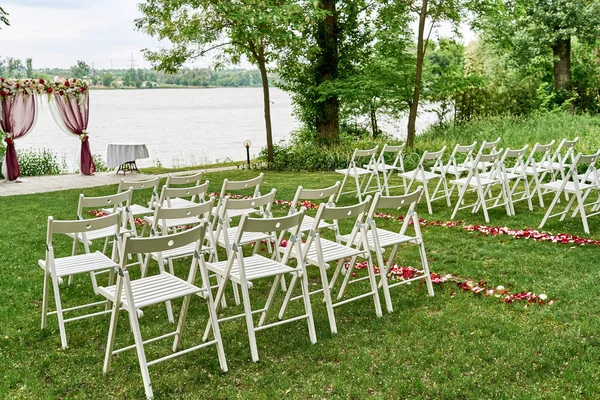 Place for wedding ceremony with wedding arch decorated with cloth, flowers and white chairs on each side of archway outdoors, copy space. Empty wooden chairs for guests on green grass. Wedding setup