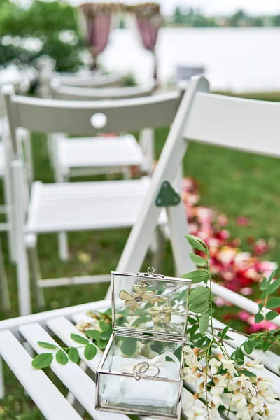 Two golden wedding rings in glass box and branch of white acacia with flowers and leaves on wooden chair. Wedding card, Valentine's Day greeting. Wedding rings, copy space