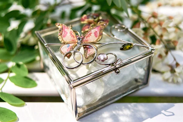 Two golden wedding rings on glass box with butterfly and branch of white acacia with flowers and leaves on wooden surface. Wedding card, Valentine\'s Day greeting. Wedding rings, copy space