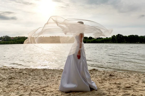 Beautiful bride in white dress standing alone on beach of lake or river outdoors, copy space. Wedding dress, wedding details. Veil fluttering in the wind