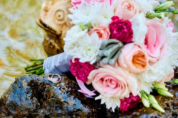 Close up of two golden wedding rings near  beautiful fresh wedding bouquet of pink and white roses in blur on stone in water, free space. Wedding details outdoor with copy space