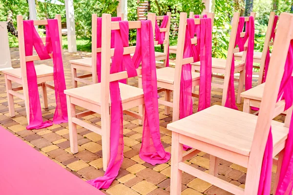 Wedding chairs on each side of archway. Place for wedding ceremony decorated in pink color, wooden chairs for guests outdoors. Wedding ceremony in pink color