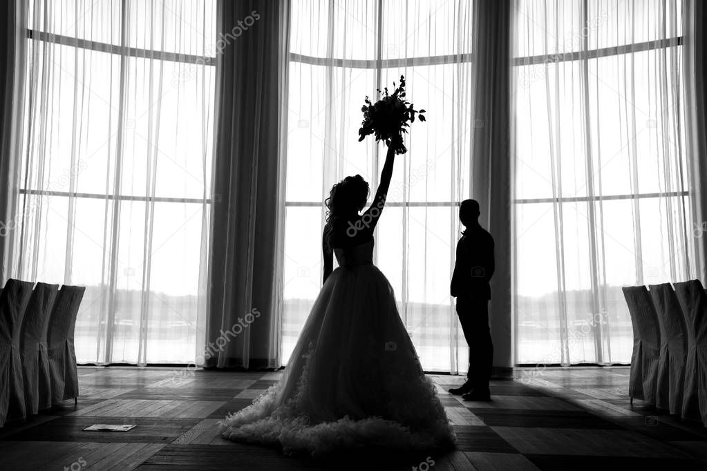 Silhouettes of bride and groom at the window indoors, free space. Bride holding wedding bouquet in her raised hand. Silhouettes of wedding couple, full length. Black and white photo