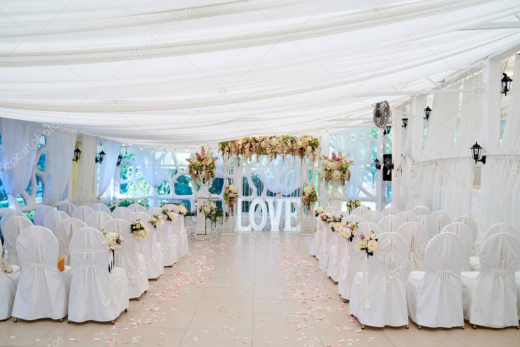 Place for wedding ceremony with white wedding arch decorated with flowers and chairs for guests on each side, copy space. Beautiful wedding archway with rose petals
