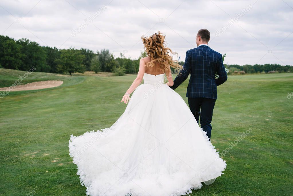 Full length body portrait of young bride and groom running on green grass of golf course, back view. Happy wedding couple walking through golf course