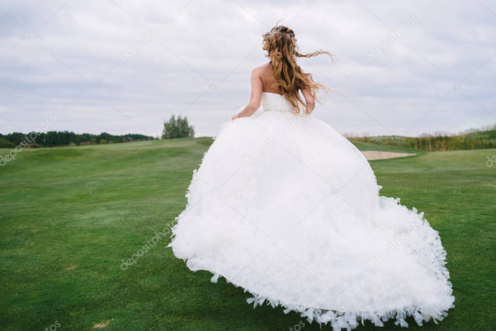 Full length body portrait of beautiful bride in fashion white wedding dress with feathers running away through green golf course, back view. Runaway bride