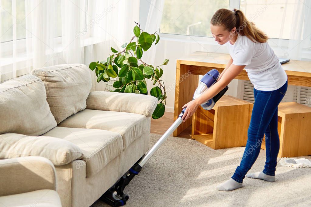 Full length body portrait of young woman in white shirt and jeans cleaning carpet with vacuum cleaner in living room, copy space. Housework, cleanig and chores concept