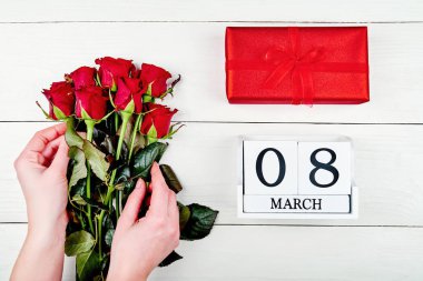 Hediye kutusu ve küp takvim yakınındaki kırmızı gül buketi ahşap arka plan üzerinde tutan kadın. Tarihi beyaz blok takvim 8 Mart kadın günler için sakla. Tebrik kartı. Üstten görünüm, düz yatıyordu