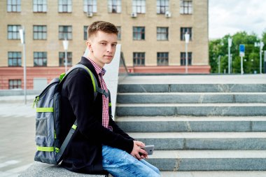 Üniversite Kampüsü açık havada ile hareket eden telefon, kopya alanı merdivenlerde oturan sırt çantası ile erkek öğrenci portresi. Eğitim, dinlenmek ve rahatlamak, seyahat ve Turizm kavramı