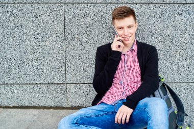 Üniversite Kampüsü, kopya alanı içinde kalan sahip tek başına gri taş duvara yakın oturmak ve cep telefonu üzerinde konuşurken gülümseyen erkek öğrenci portresi. Eğitim, dinlenmek ve rahatlamak, seyahat ve Turizm kavramı