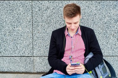Taş duvara yakın oturan ve kopya alanı cep telefonunda kulaklık, dinlenme açık havada, üniversite kampüsü içinde olması ile müzik dinlemek mutlu öğrenci. Eğitim, dinlenmek ve rahatlamak kavramı