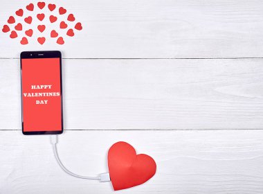 Beyaz ahşap zemin, kopya alanı üzerine kırmızı kağıt kalp ile şarj işaret mutlu sevgililer günü ile mobil akıllı telefon. Sevgililer günü için mockup şablonu. Aşk, teknoloji kavramı. Üstten görünüm, düz yatıyordu