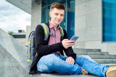 Açık havada Üniversitesi Kampüs merdivenlerde oturan ve smartphone kullanarak, yazarak, dalga köpüğü enterne, sınav için kopya alanı hazırlama sırt çantası ile gülümseyen öğrenci. Teknoloji, eğitim kavramı