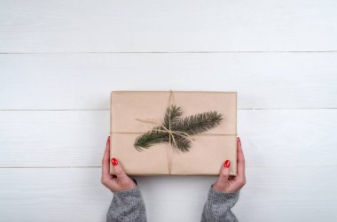 Kadın kırmızı manikür Noel hediyesi holding ile kraft kağıt beyaz ahşap masa arka plan, boş alan üzerinde sarılı. Kızın elini hediye kutusunda, yakın çekim. Noel arka plan. Düz yatıyordu, en iyi görünümü