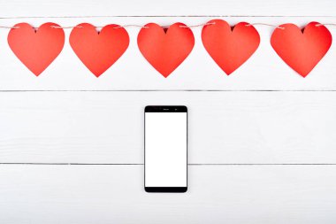 Mobil akıllı telefon ile beyaz boş perde ve kırmızı kağıt kalpler garland ahşap zemin, kopya alanı üzerine kapatın. Düz yatıyordu, en iyi görünüm. Uygulama şablonu sahte 