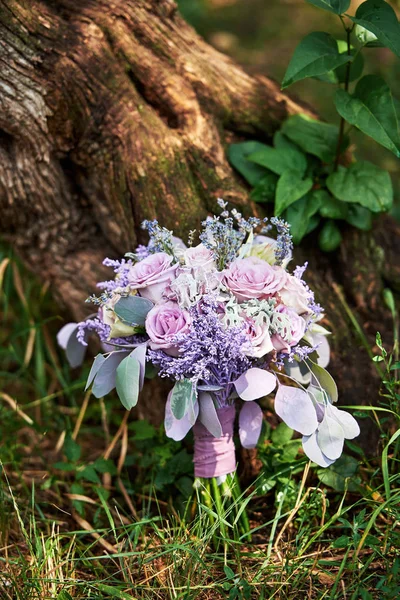 Close up of lush wedding bouquet of purple roses, violet flowers and leaves on green grass under tree, copy space. Bridal bouquet outdoors. Greeting card