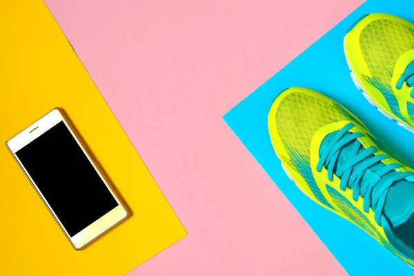 Pair of sport shoes and mobile cellphone on colorful background. New sneakers on pink, blue and yellow background, copy space. Overhead shot of running shoes and smartphone. Top view, flat lay