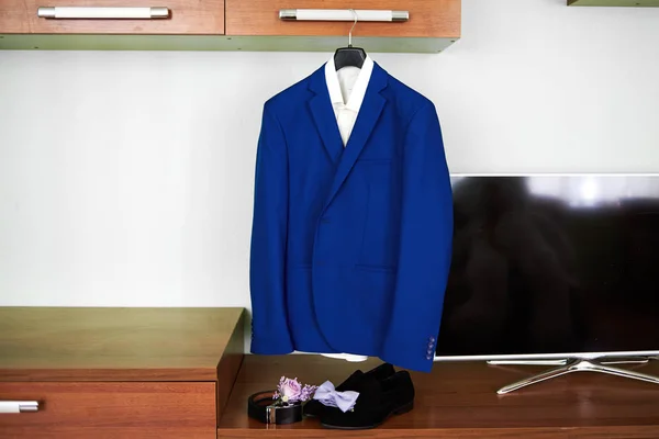 Elegant wedding blue groom suit with white shirt hanging above black suede shoes, leather belt, violet bowtie and boutonniere in room, copy space. Groom wedding accessories, free space