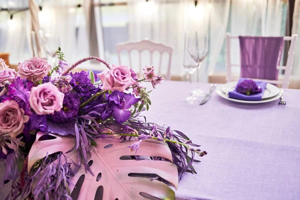 Lush floral arrangement of violet roses, purple flowers and leaves on wedding table, copy space. Wedding presidium in restaurant. Banquet table for newlyweds with flowers and violet cloth. Wedding decorations