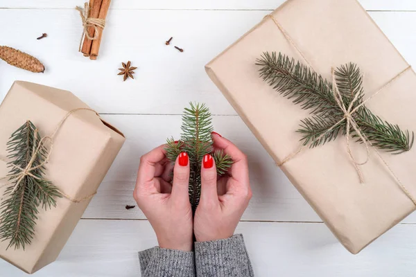 Christmas gift wrapping background, copy space. Female hands holding branch of fir tree, gift boxes wrapped in kraft, top view. Winter holidays concept, flat lay. Woman with red manicure packing gift