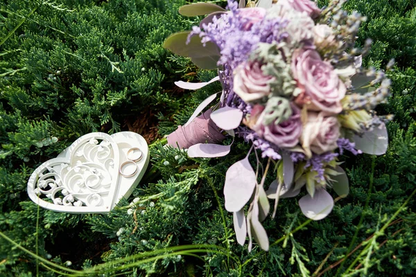 Close up of wedding rings in box in heart shape and lush wedding bouquet of purple flowers in blur on green grass background outdoors, copy space. Wedding concept