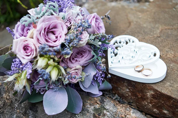 Close up of wedding bouquet of purple roses, violet flowers and leaves and wedding rings in box in heart shape on stone background, copy space. Bridal bouquet outdoors, soft focus