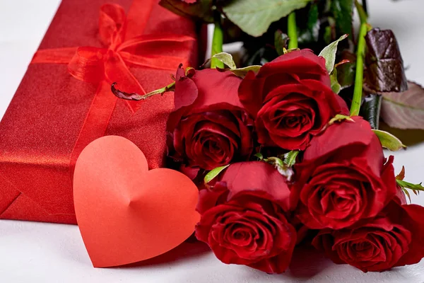 Bir buket kırmızı gül, şerit yay ile kırmızı hediye kutusu ve bir kağıt kalp Beyaz ahşap zemin üzerine kapatın. Kadının gün, Sevgililer günü, anneler günü (Mart 8 için tebrik kartı)