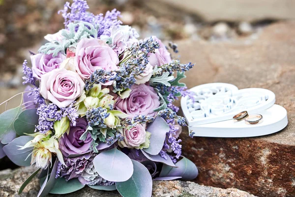 Close up of lush wedding bouquet of purple roses, violet flowers and leaves and wedding rings in box in heart shape on stone background, copy space. Bridal bouquet outdoors. Wedding concept