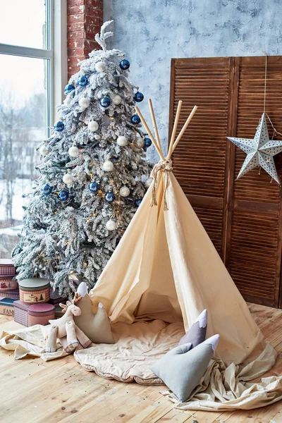 Christmas tree with gifts and wigwam near window in child room, copy space. Childen room interior with decorated play tipi tent. Scandinavian style