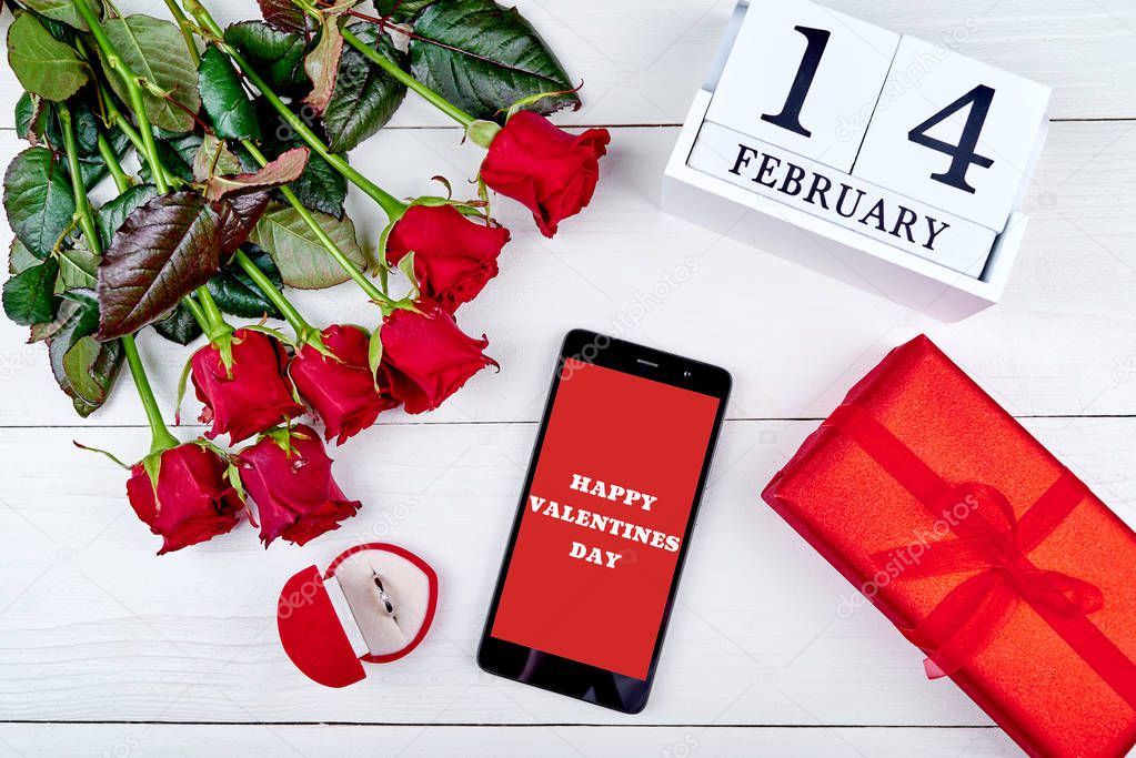 Bouquet of red roses, cellphone with sign Happy Valentines day, diamond ring, gift box and february 14 wooden calendar, copy space. Greeting card mockup. Love concept. Top view, flat lay
