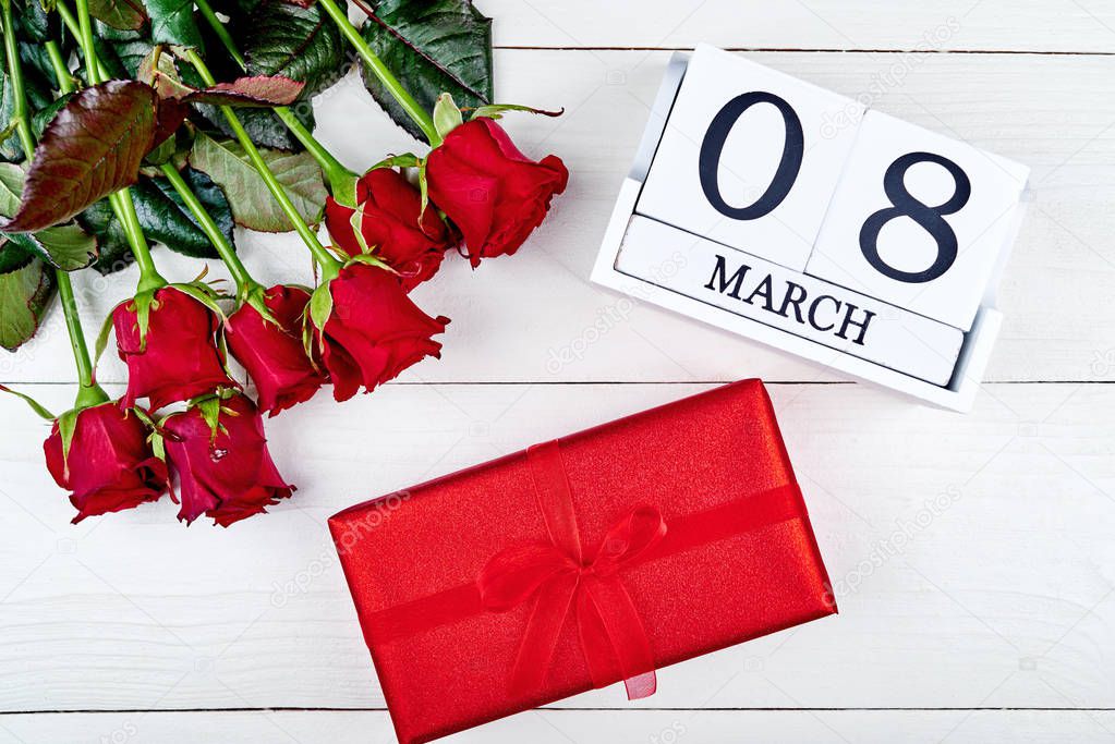 Red gift box, roses and cube calendar on white wooden background, copy space for text. Save the date white block calendar for Womens Day, March 8. Greeting card. Top view, flat lay