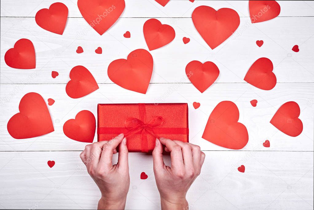 Woman holding and unwrapping red gift box on table with red hearts. Greeting card for Valentins Day, Womens Day, March 8, Mothers Day, birthday, wedding. Top view, flat lay