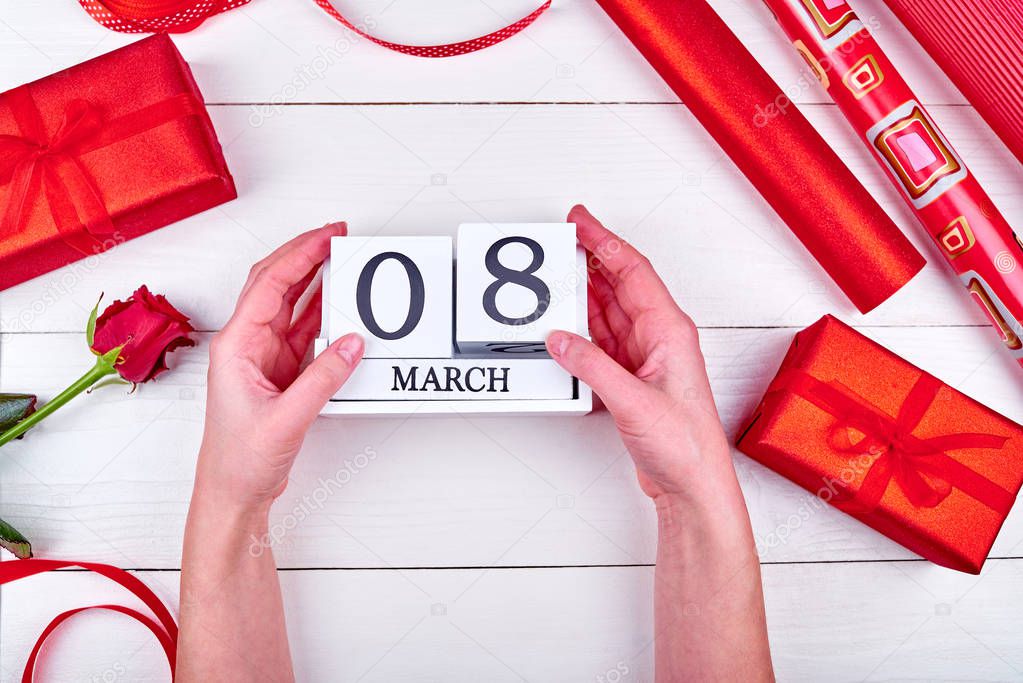 Womens Day background, copy space. Rolls of red wrapping paper, ribbon, red rose, gift boxes on table. Woman holding wooden block calendar with date March 8. Flat lay, top view 