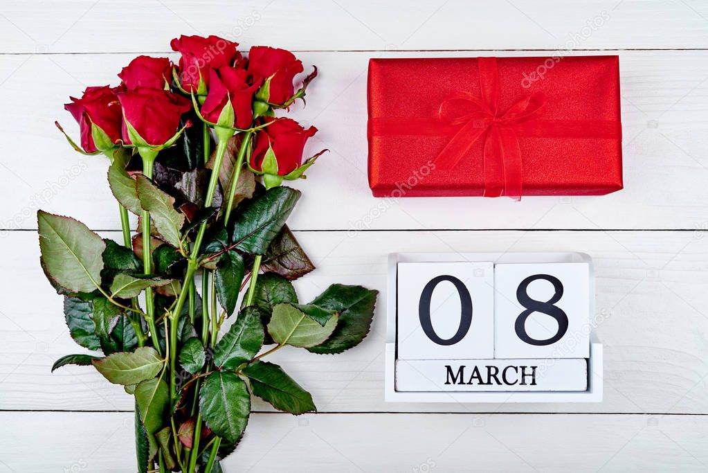 Bouquet of red roses, gift box and cube calendar on white wooden background, copy space for text. Save the date white block calendar for Womens Day, March 8. Greeting card. Top view, flat lay