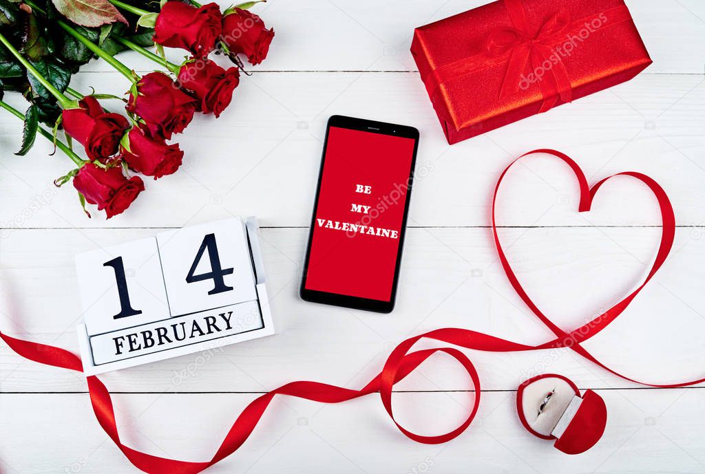 Bouquet of red roses, ribbon shaped as heart, cellphone with sign Be my valentine, diamond ring, gift box and february 14 wooden calendar, copy space. Greeting card mockup. Top view, flat lay