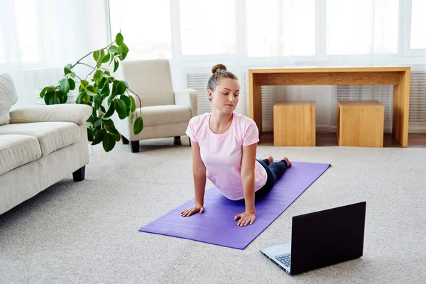 Young woman practicing yoga with laptop at home indoor, copy space. Girl practicing cobra pose, full length. Relaxing and doing yoga. Wellness and healthy lifestyle. Bhujangasana