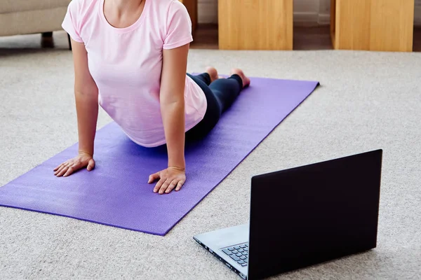 Young woman practicing yoga with laptop at home indoor, copy space. Girl practicing cobra pose. Relaxing and doing yoga. Wellness and healthy lifestyle. Bhujangasana