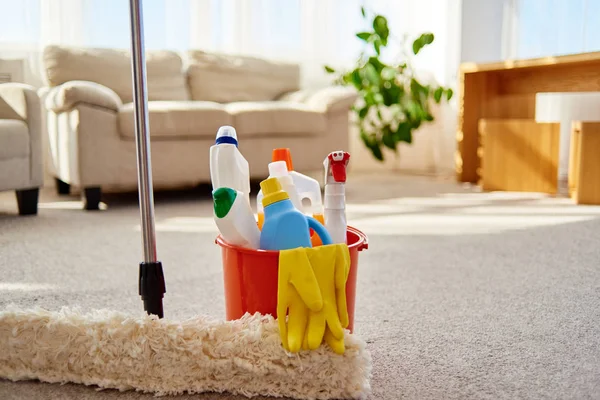 Cleaning set for different surfaces in orange bucket and mop on floor in living room, copy space. Cleaning service concept. Bucket with cleaning items for kitchen, bathroom or toilet